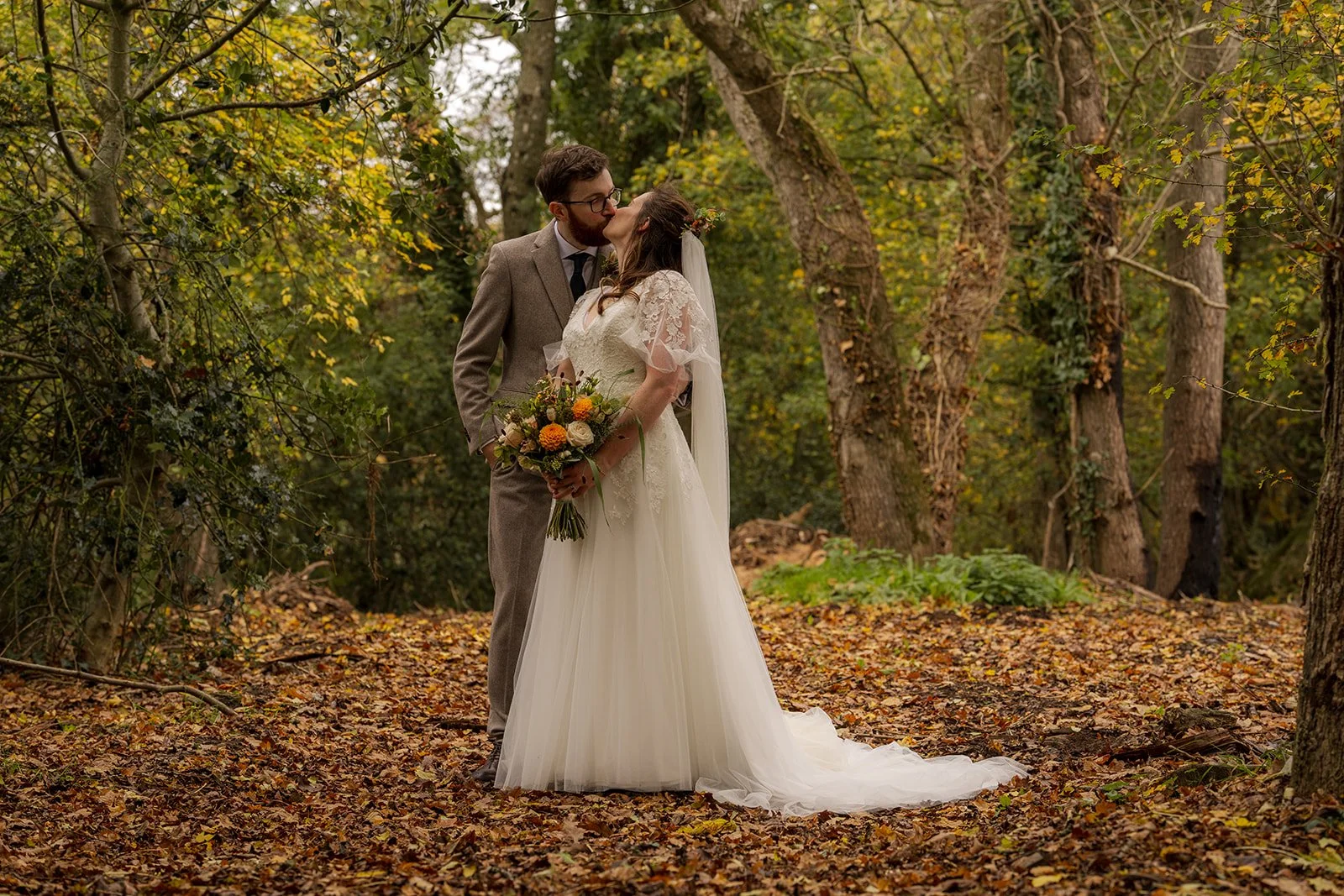 Maisie & Andrew kissing in autumn woodland at Stockbridge Farm Barn in Dorset, surrounded by golden leaves, photographed by Just James Weddings for romantic seasonal wedding portraits.