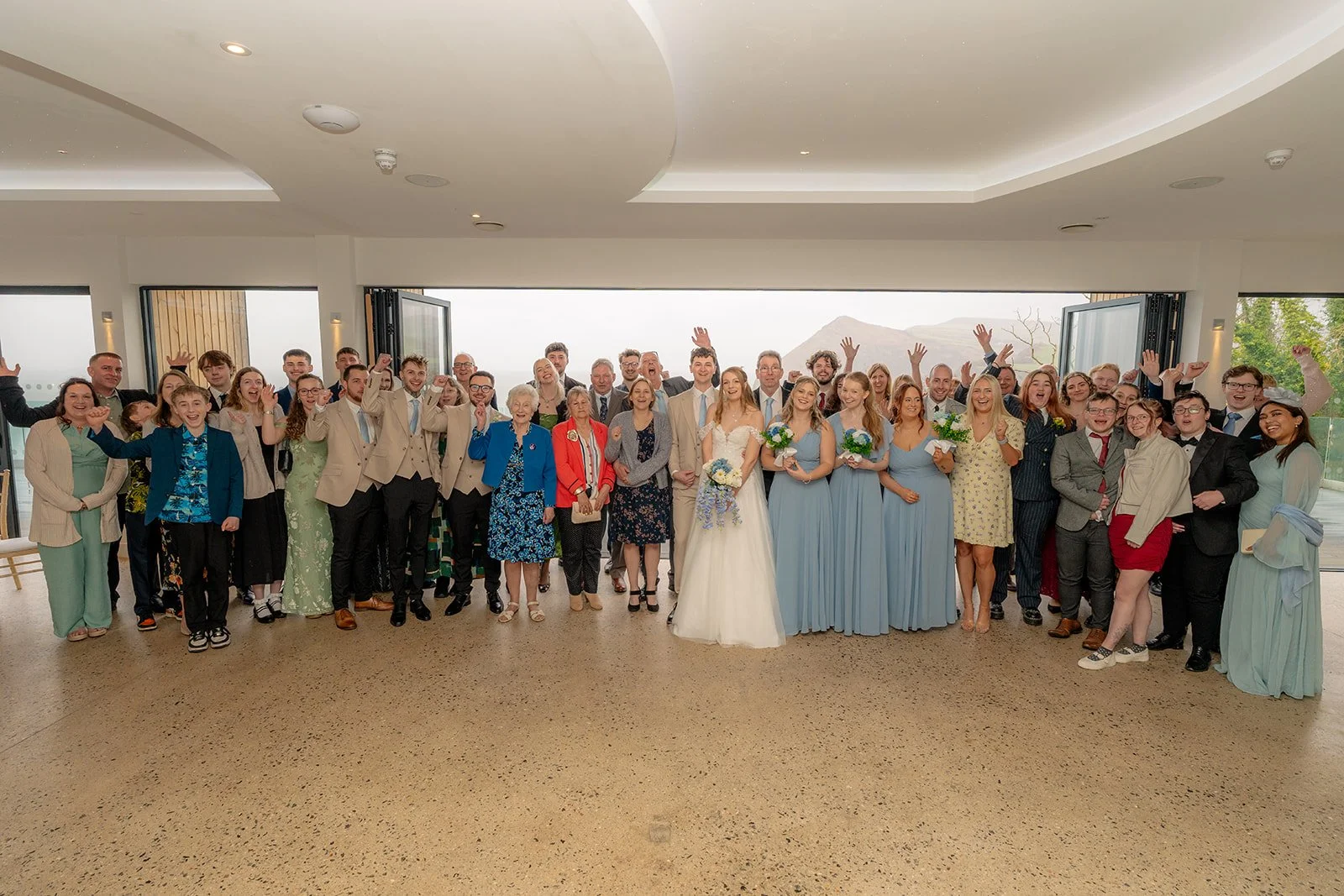 Hannah & Cameron celebrate with family and friends during a joyful group photo at Sandy Cove Hotel, North Devon. Wedding guests gather around the couple and wedding party inside the venue with coastal views behind them.