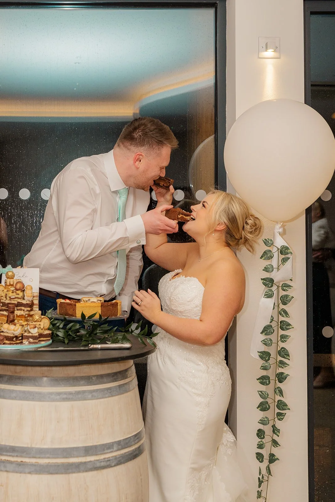 Andrew & Daisy sharing cake together at Sandy Cove Hotel in Devon, laughing as they feed each other dessert beside a barrel cake table and balloon décor, capturing relaxed evening reception moments at this seaside wedding venue.