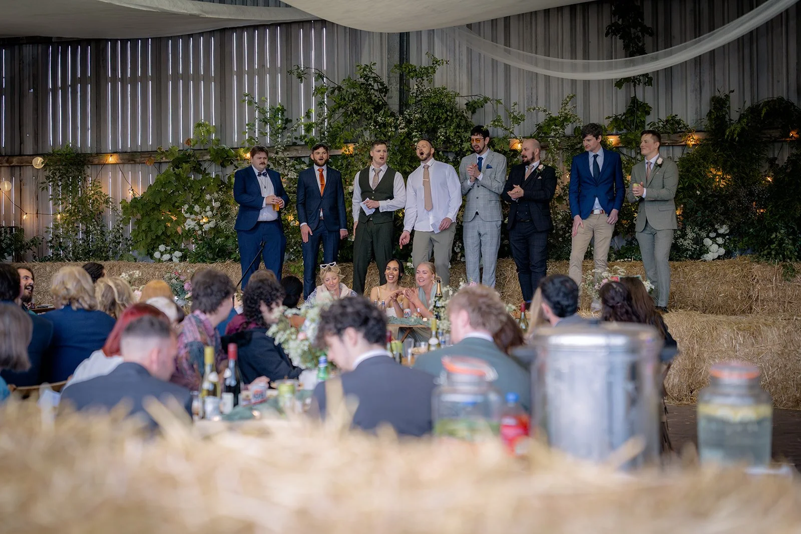 Jaye & Josh's Farrington's Farm wedding in Bristol with the groomsmen leading a lively sea shanty-style moment on hay bales, surrounded by cheering guests, greenery, soft draped fabric and warm barn reception atmosphere full of personality.
