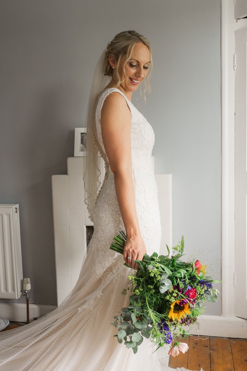 A bride in a white lace wedding dress and veil holding a colorful bouquet of flowers, standing indoors with a light gray wall behind her.