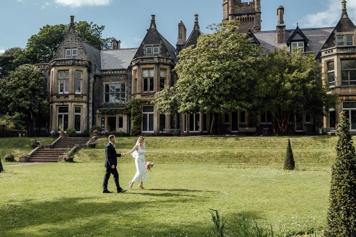 A bride and groom holding hands in a garden with a large historic mansion in the background.