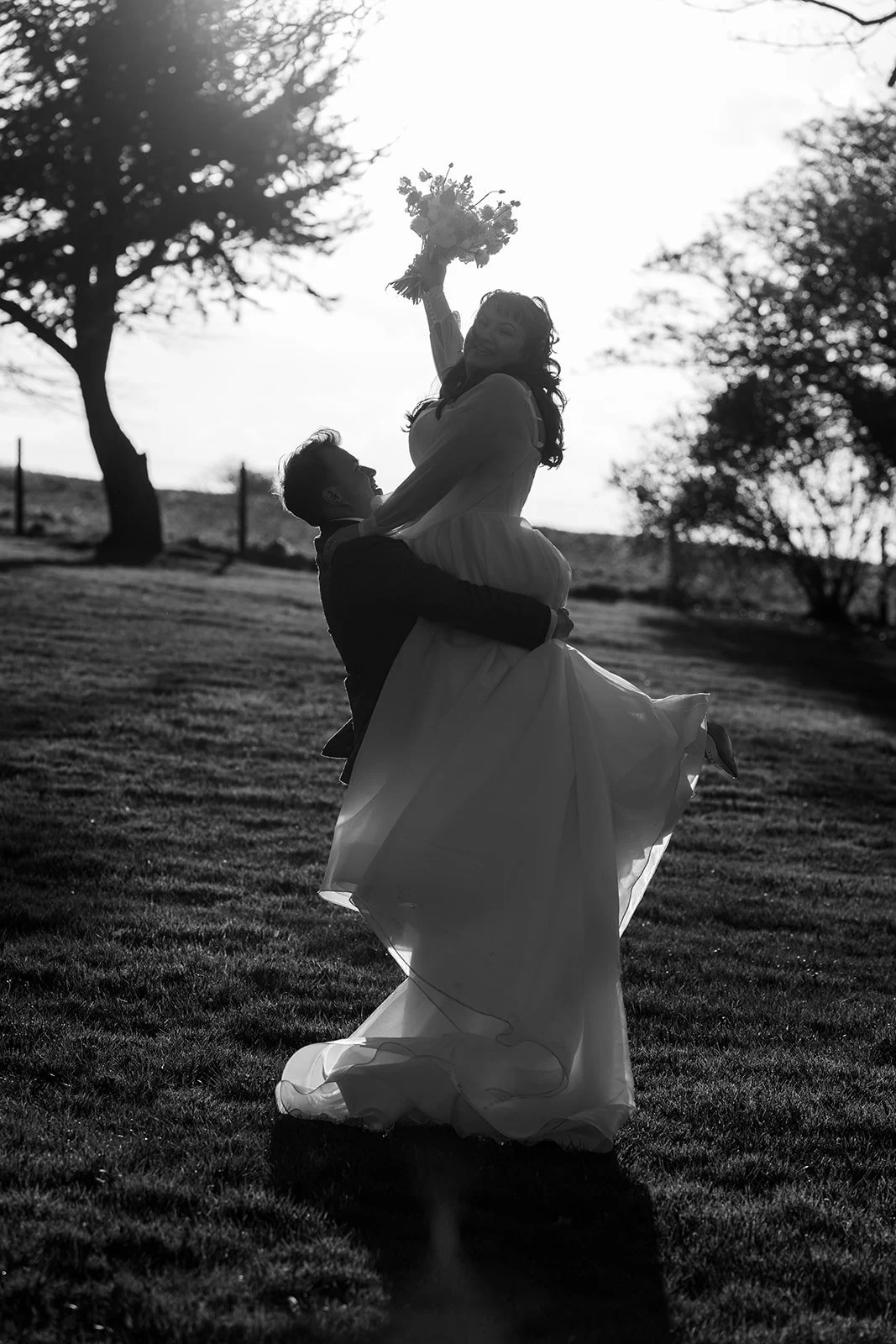 Emily & Josh on the lawns at Coombe Lodge Blagdon, Somerset, UK, as Josh lifts Emily into the air and she raises her bouquet overhead. Outdoor wedding photo in silhouette with trees, evening light and the bride’s dress flowing.