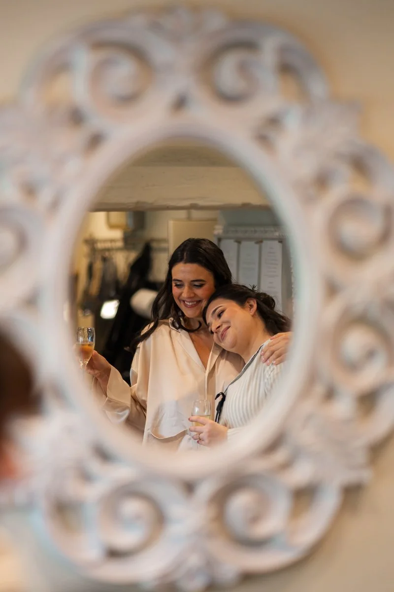 Megan &amp; Luca wedding morning reflected in an ornate mirror at The Great Barn, Devon, showing two smiling bridesmaids with champagne sharing a quiet, joyful moment. Candid wedding prep photography with soft light and romantic documentary detail. 