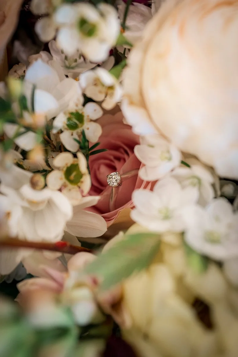  Close-up wedding ring photo from Megan &amp; Luca’s day at The Great Barn, Devon, with the band nestled among creamy roses, blush petals and delicate white flowers. Romantic macro wedding photography with floral detail and soft, elegant texture. 