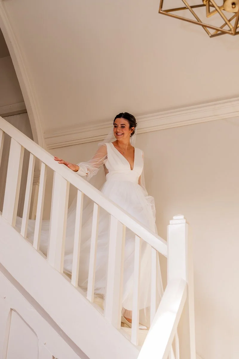  Bride Megan walks down a white staircase in soft window light at The Great Barn, Devon, wearing a long-sleeved wedding dress and smiling during morning preparations. Elegant bridal portrait photography from Megan &amp; Luca’s wedding in Devon, UK. 