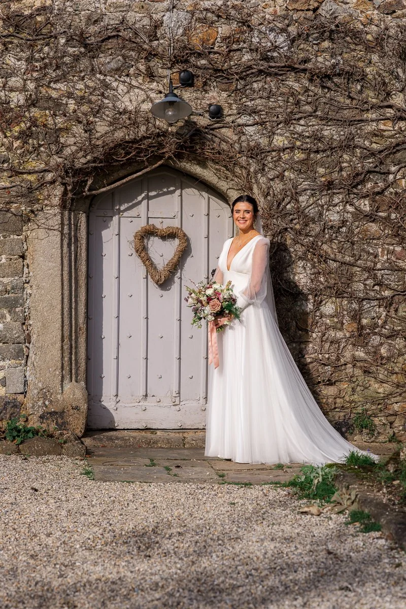  Bride Megan stands by a rustic stone doorway with a heart wreath at The Great Barn, Devon, holding her bouquet in soft sunlight. Romantic bridal portrait photography capturing Megan &amp; Luca’s wedding style against the barn’s charming countryside 