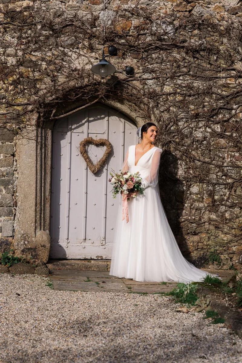  Bride Megan poses beside a weathered stone wall and heart-adorned doorway at The Great Barn, Devon, holding a pastel wedding bouquet in golden light. Timeless bridal portrait from Megan &amp; Luca’s wedding, capturing elegant Devon barn wedding phot