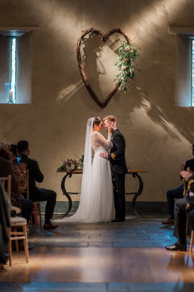  Megan &amp; Luca stand together during their ceremony at The Great Barn, Devon, beneath a rustic heart arrangement on the chapel wall. Soft light, intimate framing and elegant styling capture a romantic barn wedding moment in this Devon wedding phot