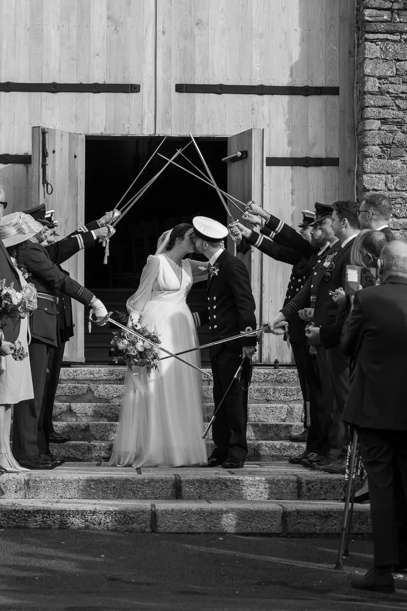  Black and white wedding photo of Megan &amp; Luca kissing beneath a sword arch outside The Great Barn, Devon, surrounded by guests and naval uniforms. A timeless confetti-style exit moment capturing military wedding tradition and Devon barn wedding 