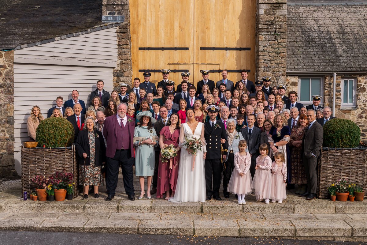  Megan &amp; Luca pose with their full wedding party and guests outside The Great Barn, Devon, in a classic group portrait after the ceremony. This joyful Devon barn wedding photo captures family, friends and military detail in countryside light. 