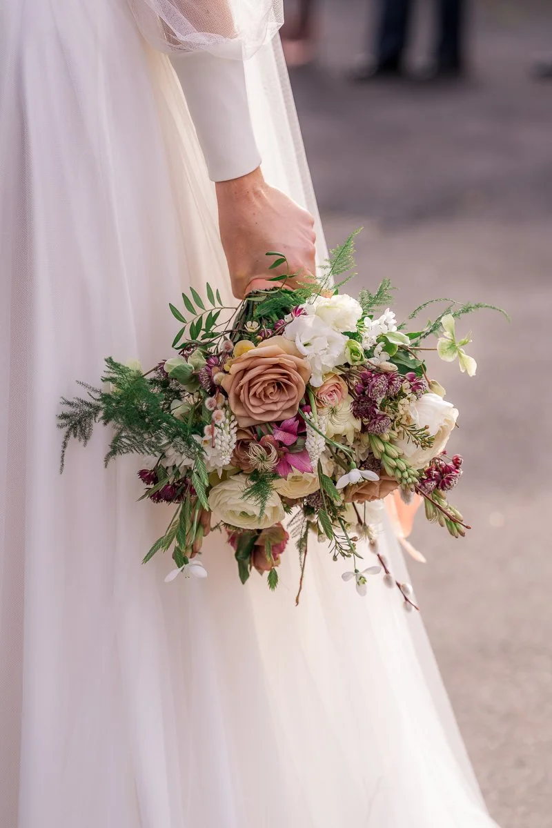  Megan’s bouquet is held against her wedding dress at The Great Barn, Devon, featuring blush roses, ivory blooms and trailing greenery. Elegant floral detail from Megan &amp; Luca’s wedding, ideal for Devon wedding inspiration and barn wedding. 