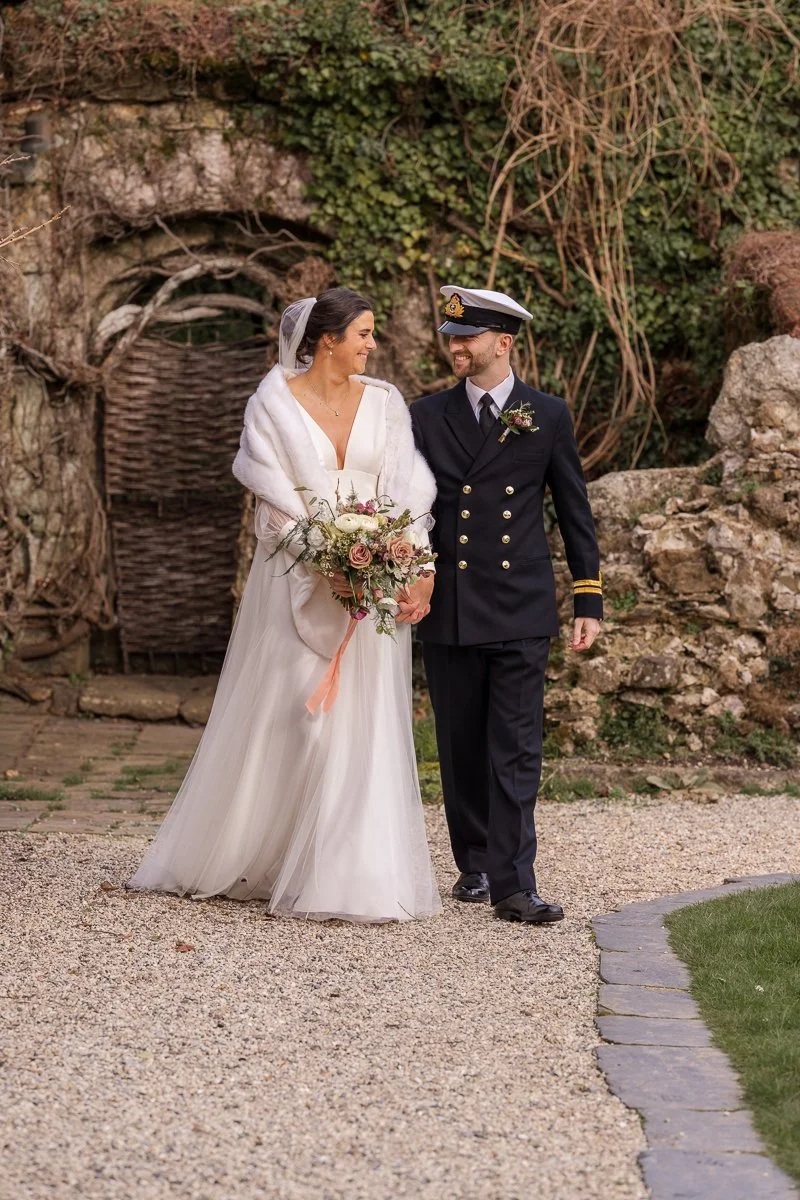  Megan &amp; Luca walk together through the gardens at The Great Barn, Devon, with Megan holding her bouquet and Luca in naval uniform. Romantic Devon barn wedding photography captures a relaxed just-married moment in soft countryside evening light. 