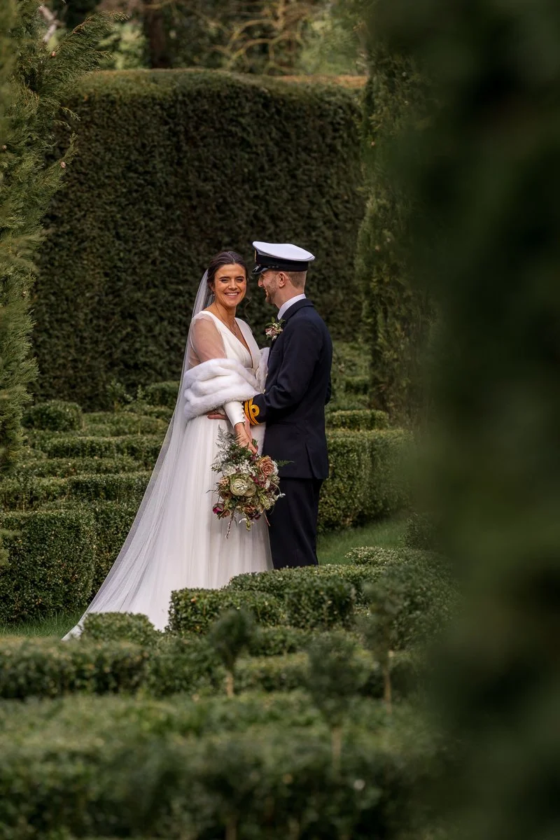  Megan &amp; Luca stand together in the formal gardens at The Great Barn, Devon, framed by tall hedges and clipped greenery. This romantic wedding portrait captures the couple in soft light, with Luca in naval uniform and Megan holding her bouquet. 