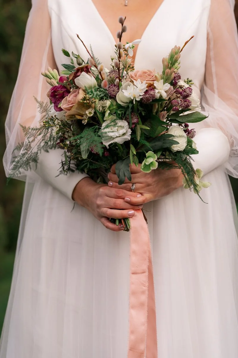  Megan holds her bouquet against her wedding dress at The Great Barn, Devon, showing blush roses, soft ivory blooms and trailing greenery. Elegant bridal floral detail from Megan &amp; Luca’s wedding, perfect for Devon barn wedding inspiration and SE