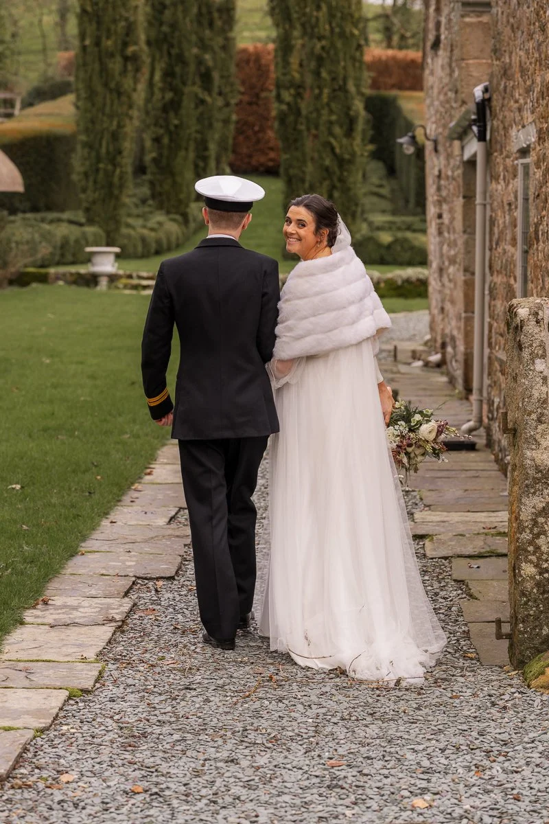  Walking hand in hand beside The Great Barn, Devon, Megan looks back smiling at the camera while Luca leads her along the garden path. This romantic wedding portrait captures bridal style, naval uniform detail and the venue’s countryside setting. 