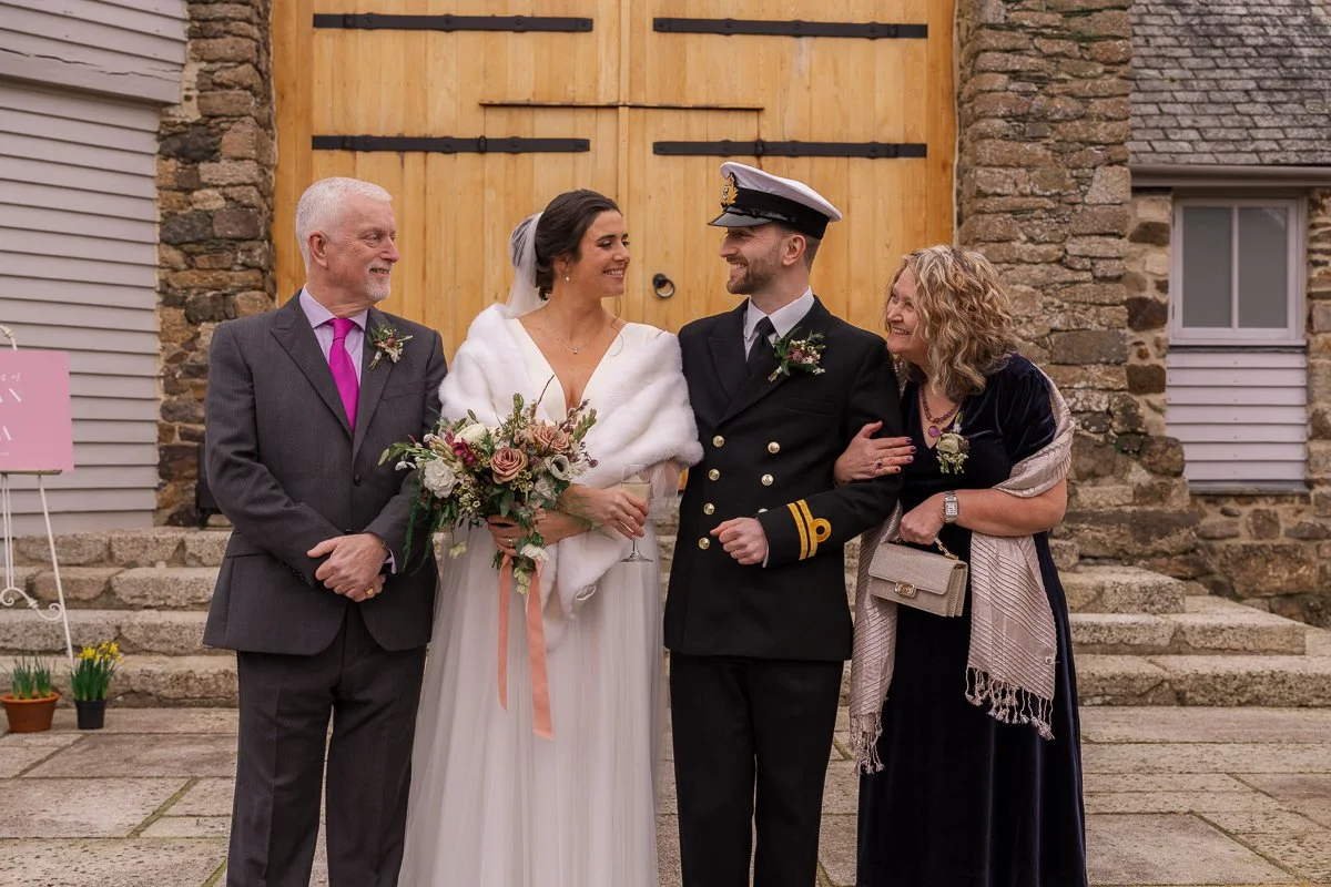  Megan &amp; Luca stand outside The Great Barn, Devon, with family after their wedding ceremony, smiling in front of the barn doors as Megan holds her bouquet. Natural family wedding photography capturing a warm, candid moment at this Devon barn wedd