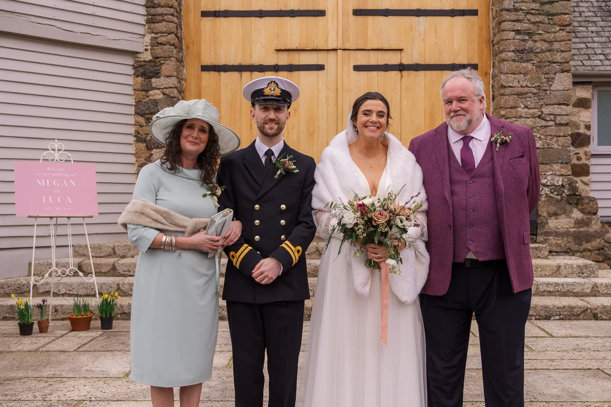  Megan &amp; Luca pose with family outside The Great Barn, Devon, in front of the barn doors after their ceremony, with Megan holding her bouquet and Luca in naval uniform. Classic family wedding portrait photography from their elegant Devon barn wed