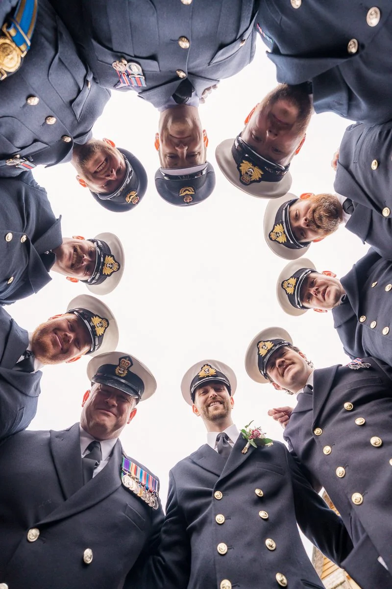  Naval officers gather in a circle around groom Luca at Megan &amp; Luca’s wedding at The Great Barn, Devon, looking down into the lens in a playful group portrait. Fun military wedding photography with crisp uniform detail at this Devon venue, UK. 
