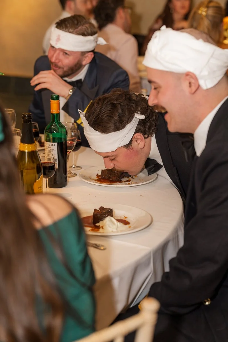  Guests in playful napkin hats laugh during the wedding meal at The Great Barn, Devon, as one friend leans into his dessert at Megan &amp; Luca’s celebration. Candid reception photography capturing fun, humour, warm party atmosphere in Devon, UK. 