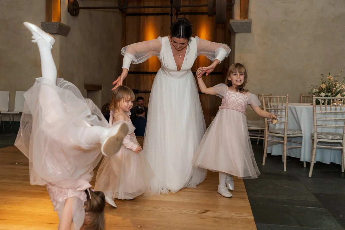  Megan dances with two young flower girls at The Great Barn, Devon, while another child cartwheels across the dance floor beside them. Joyful wedding reception photography from Megan &amp; Luca’s day, full of movement, fun and family energy in Devon.