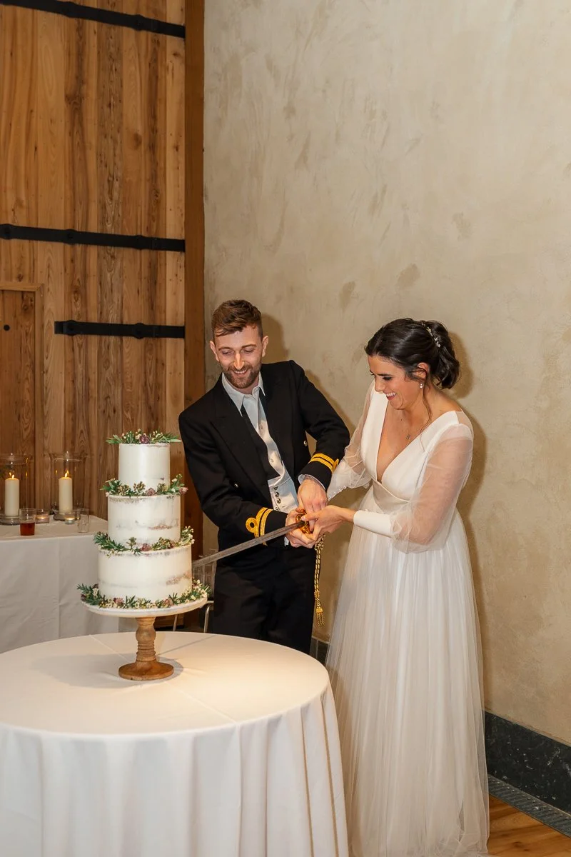  Megan &amp; Luca cut their wedding cake at The Great Barn, Devon, standing beside a three-tier floral cake against the barn’s rustic interior wall. Joyful reception photography captures this classic wedding moment with elegant styling at their Devon