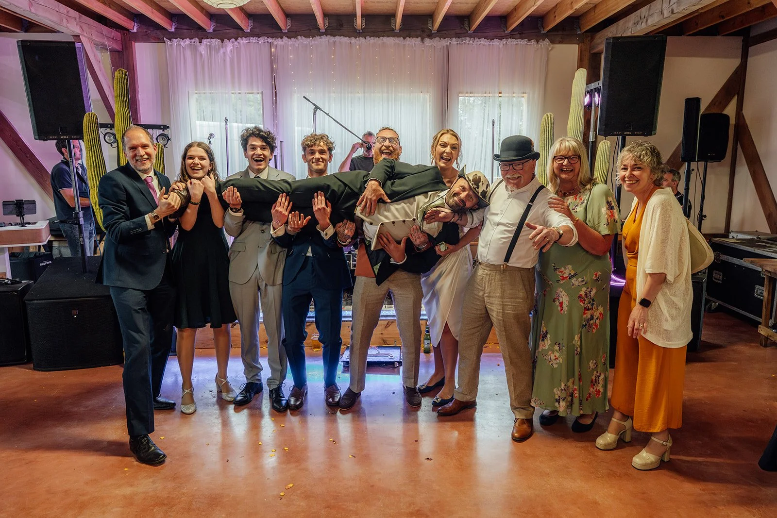 Fun reception group photo from Hannah & Ben’s wedding at Grays Barn in Devon, UK, with guests laughing as Ben is lifted across the dance floor in front of the band and stage lights, captured with lively party energy by Just James Weddings.
