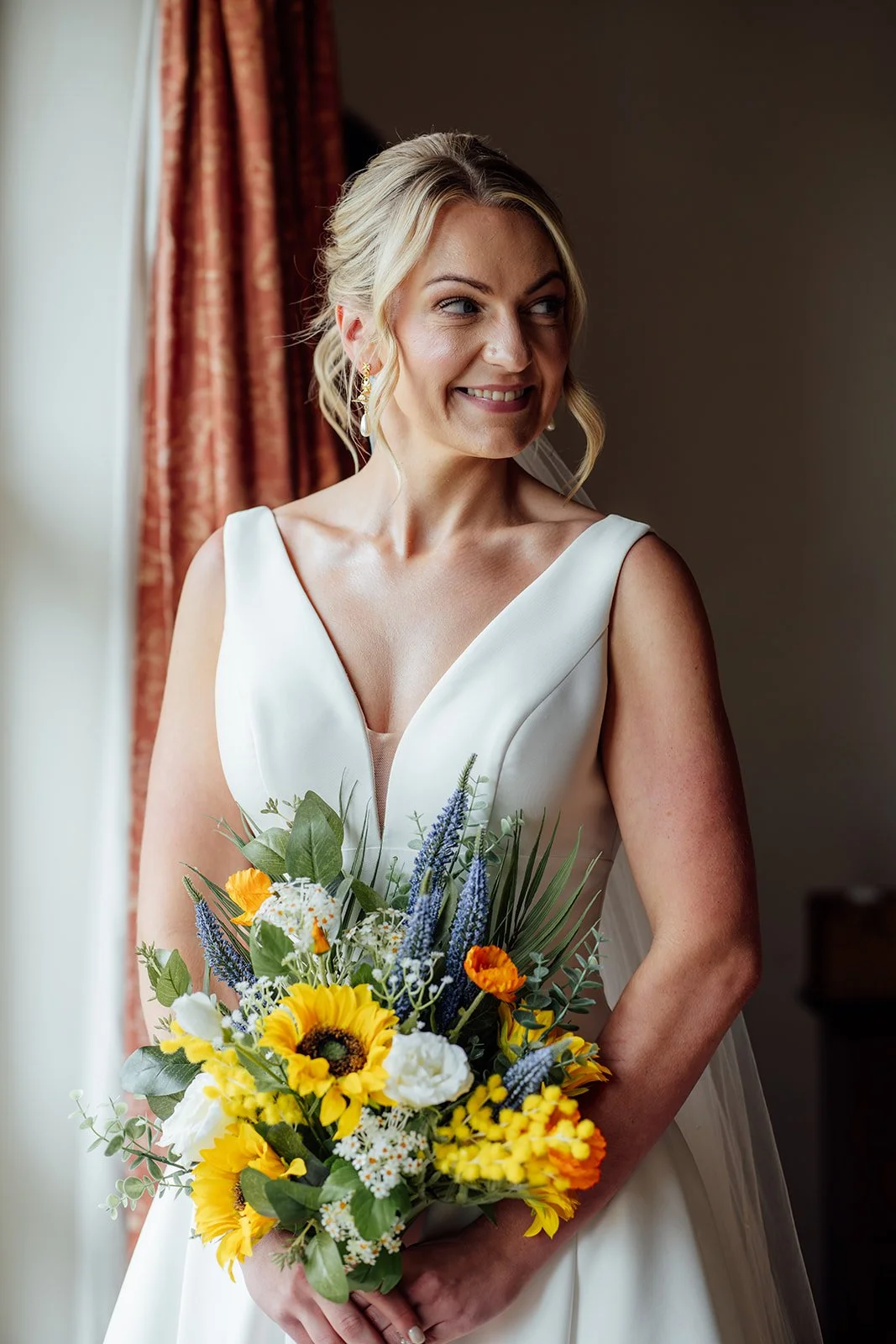 Portrait of Hannah holding a bright sunflower wedding bouquet at Grays Barn in Devon, UK, wearing her white bridal gown and pearl earrings by the window during calm, elegant morning preparations, photographed beautifully by Just James Weddings.
