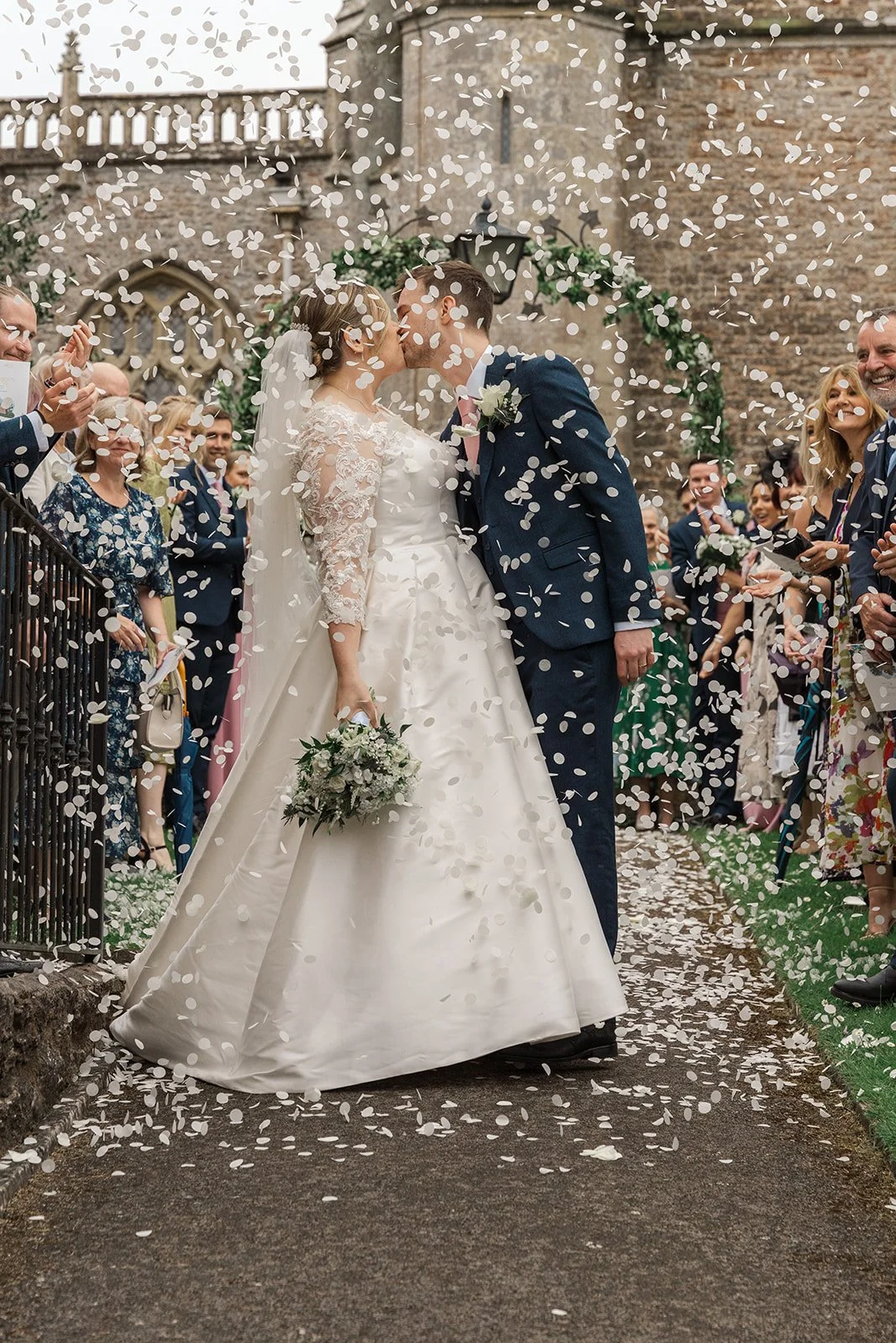 Beth & Tom kiss during their confetti exit at Bishop’s Palace in Somerset, UK, surrounded by cheering wedding guests and fluttering white petals, with the historic stone architecture framing a romantic outdoor wedding portrait.