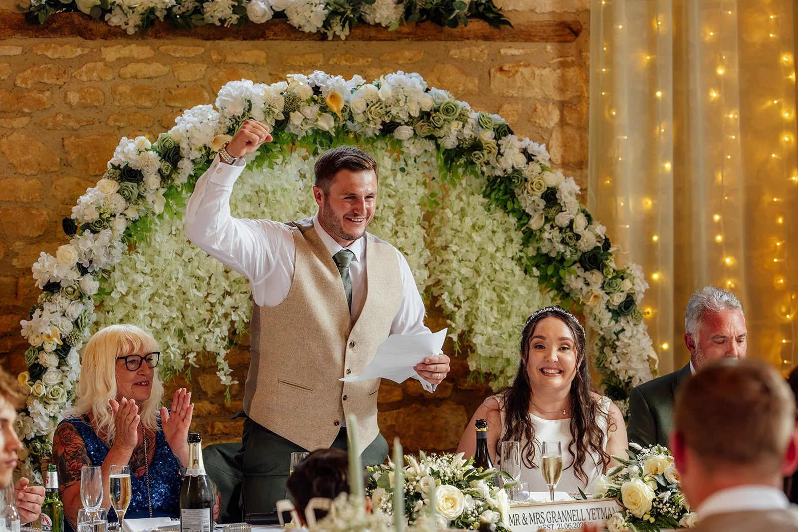Armani giving a wedding speech beside Chloe at The Barn at Upcote in the Cotswolds, with a floral backdrop and guests around them, photographed by Just James Weddings for candid reception moments.