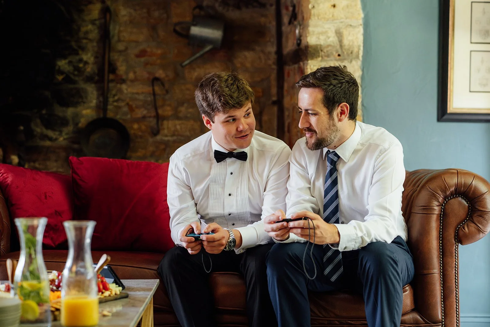 Tom and his groomsmen sit together on a leather sofa during wedding preparations at Brympton House in Somerset, holding game controllers and smiling at each other, photographed by Just James Weddings.