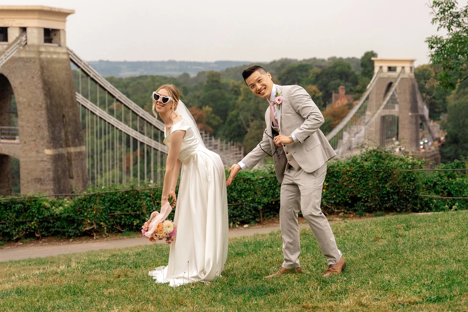 Brian & Josie posing playfully at Clifton Observatory in Somerset, with the Clifton Suspension Bridge behind them, photographed by Just James Weddings for fun relaxed wedding portraits.