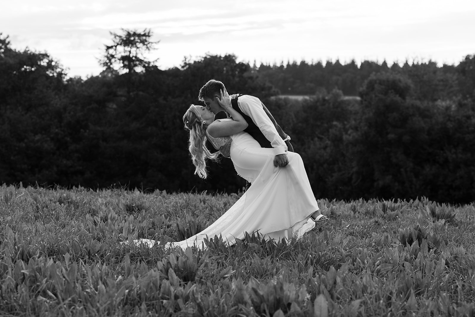 Bride and groom share a romantic dip kiss in a grassy field, her long train flowing across the ground as dark trees frame the background in a dramatic black and white portrait. Abi & Josh at The Cinder Barn celebrate just married moments outdoors.