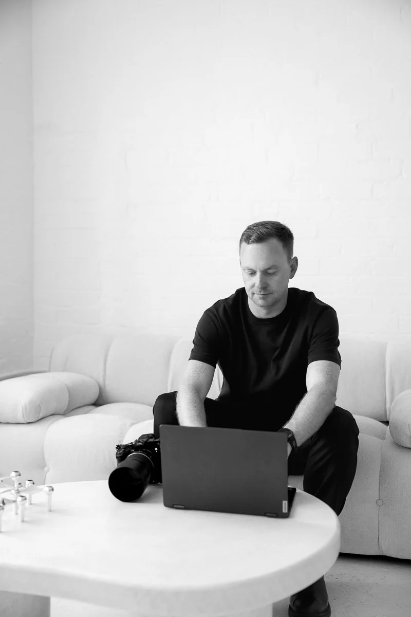 A man sitting on a sofa in a minimalistic room, working on a laptop with a camera and camera lens on the table in front of him.