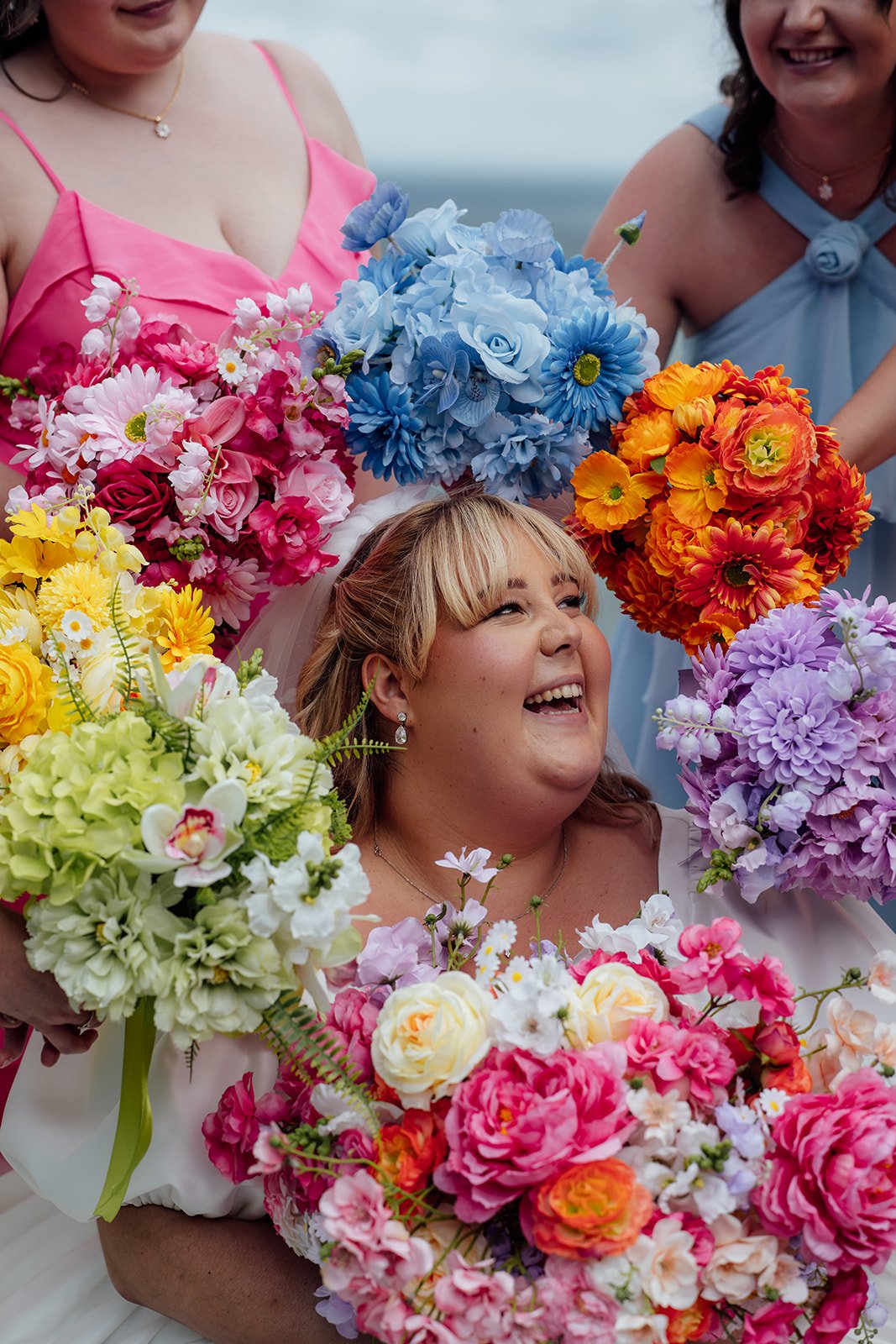 Close-up of Holly laughing among her bridesmaids’ colourful bouquets in pink, blue, orange, yellow and lilac at Sandy Cove Hotel in North Devon, photographed by Just James Weddings.