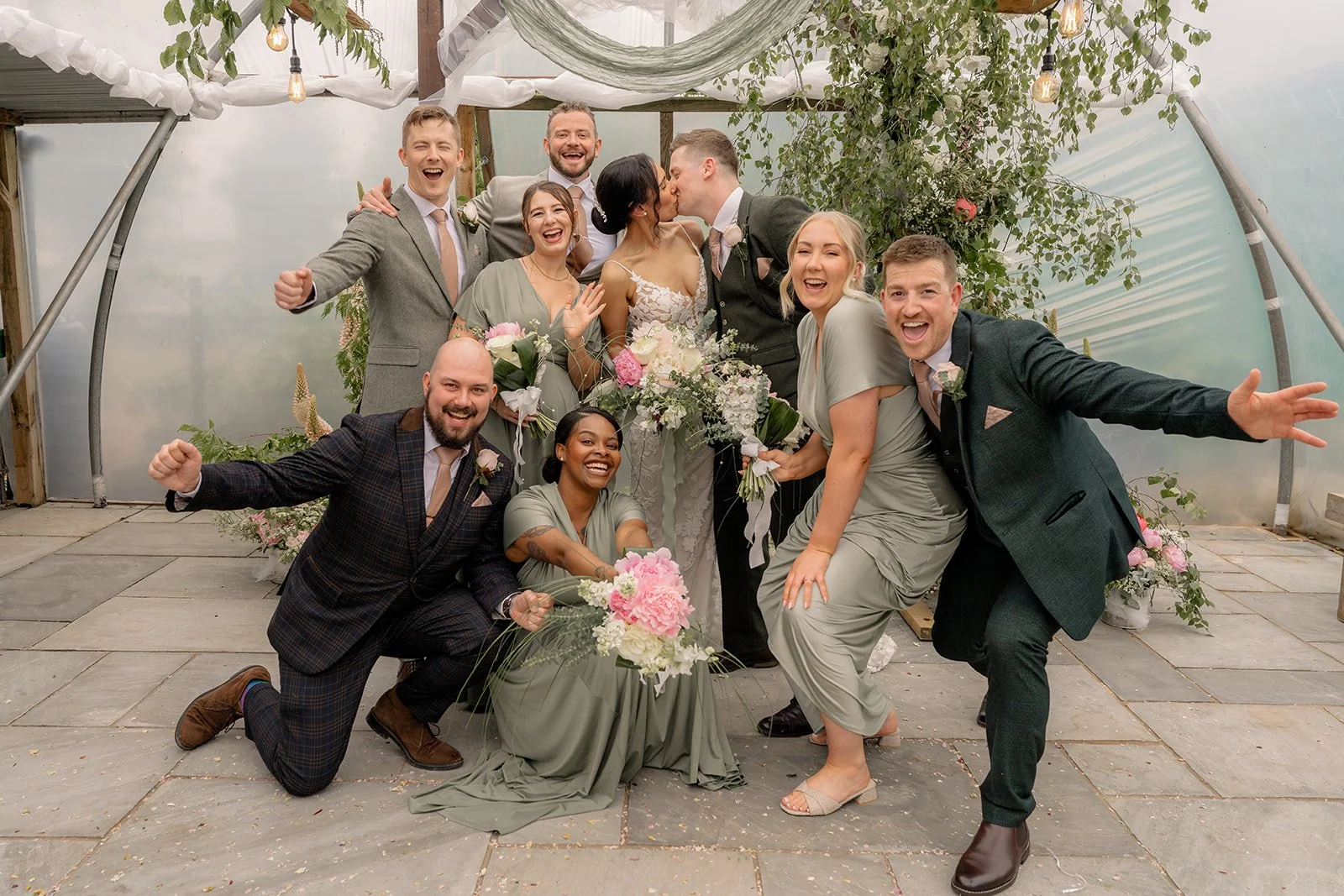 Jade and Greg kissing in the centre of their cheering wedding party during a fun group photo at Farrington's Farm in Bristol, photographed by Just James Weddings.
