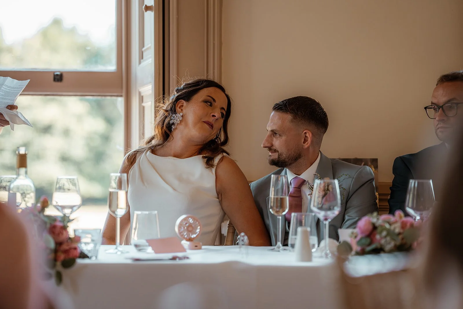 Michelle and Andy smile at each other during their wedding reception at Crowcombe Court in Somerset, UK, seated together at the top table among flowers and glassware.