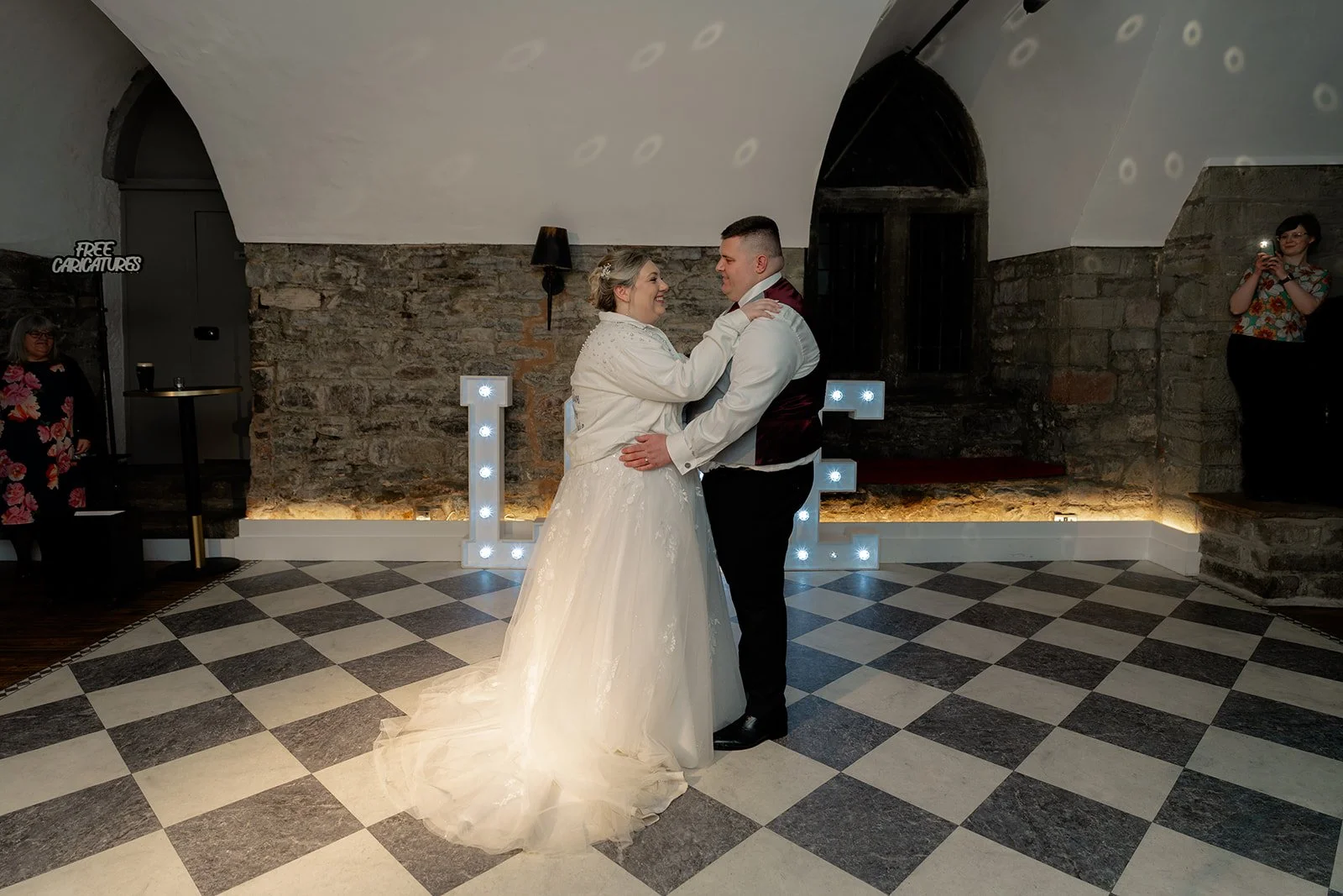 Becky & Rob share their first dance on the chequered floor during their evening reception at Clearwell Castle in Gloucestershire, UK. Romantic moment under soft lighting captured during their Clearwell Castle wedding celebration.