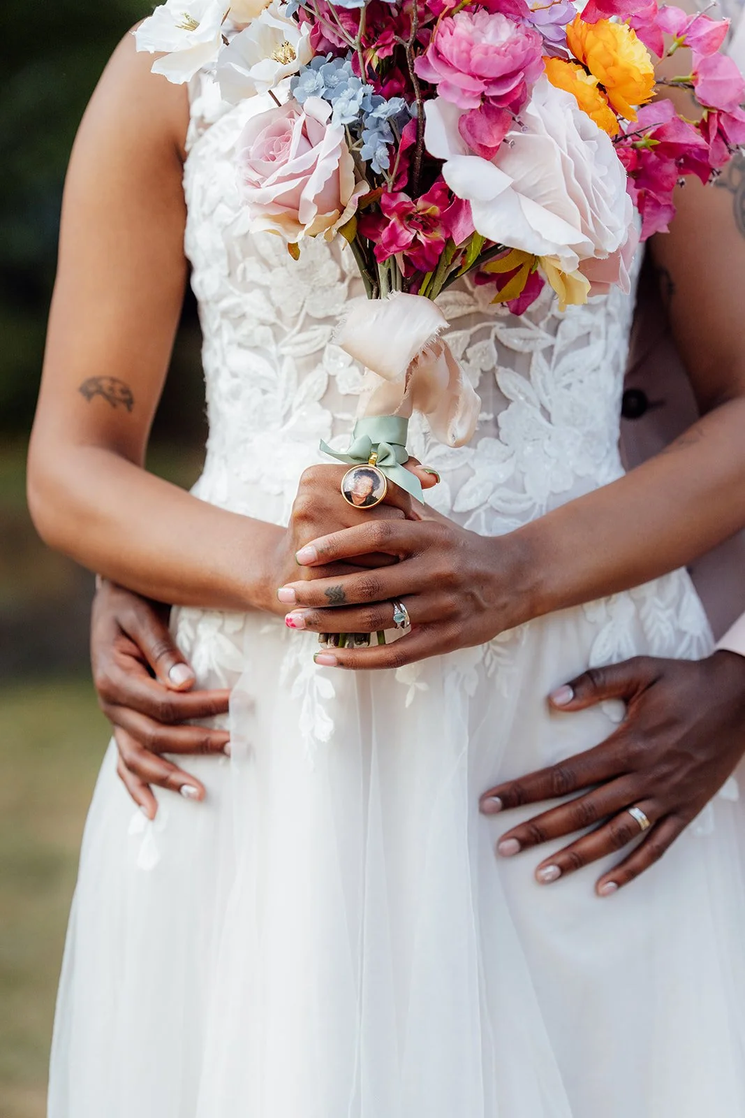 Close-up of Larisha holding her colourful bridal bouquet with a sentimental photo charm, while Kasey embraces her at Oaks Farm Weddings in Surrey.