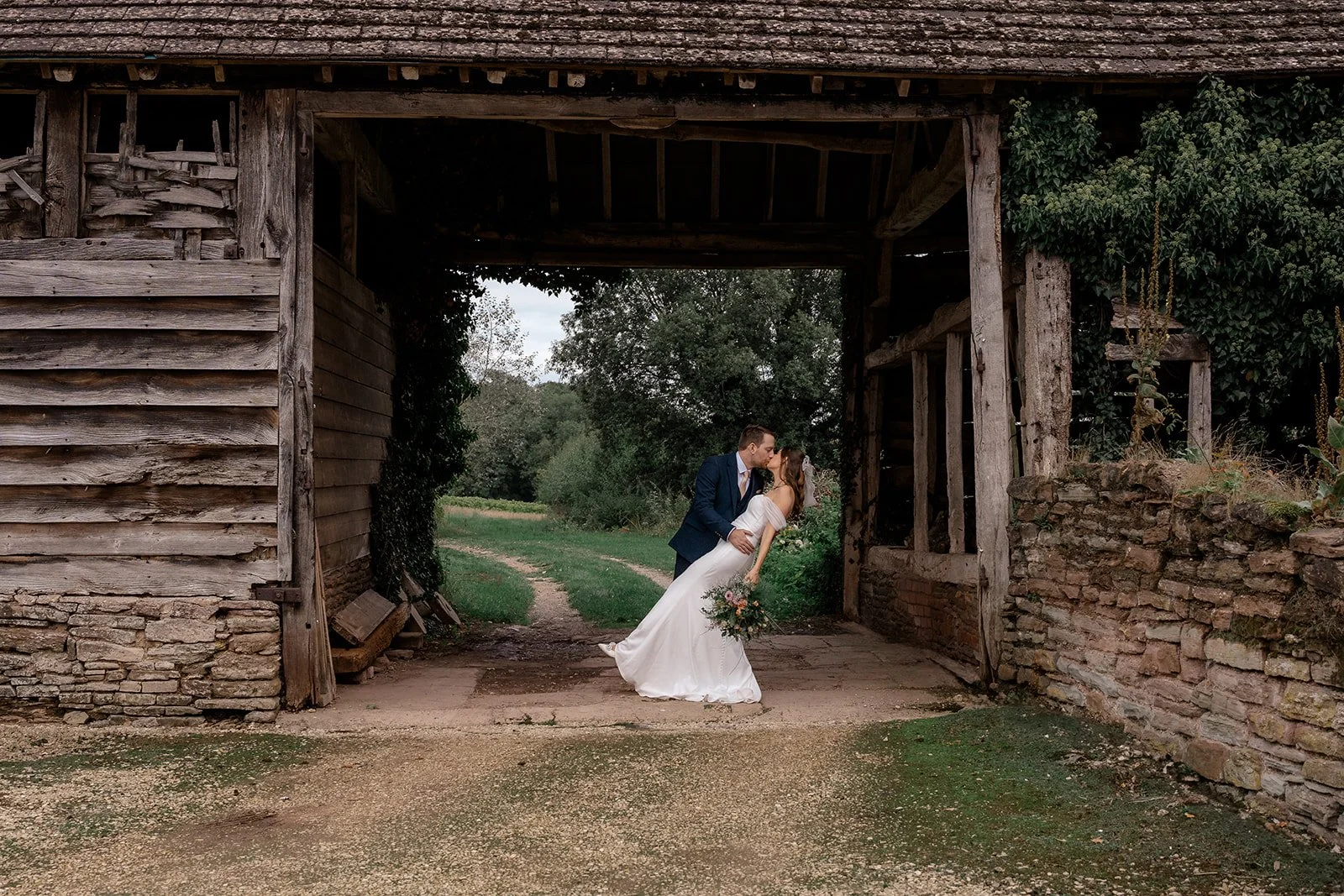 Lilly & Lloyd kissing beneath a rustic timber archway at Broadfield Court in Herefordshire, photographed by Just James Weddings for relaxed countryside wedding portraits.