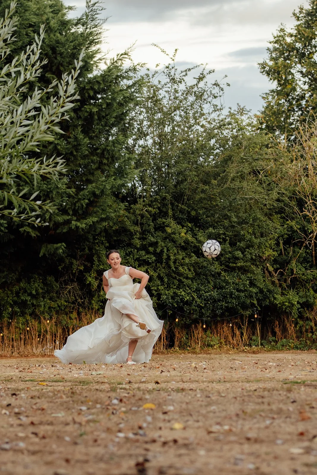 Harriet kicks a football across the grounds in her wedding dress during a playful moment at Dovecote Events in Oxfordshire, photographed by Just James Weddings.