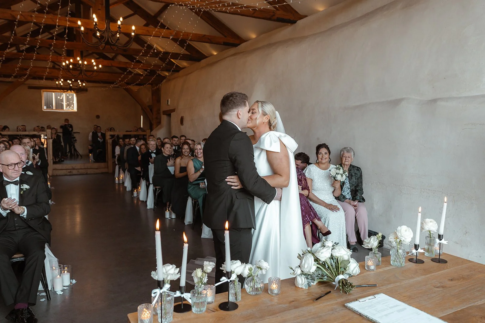 Jessie & Jay share their first kiss during the wedding ceremony at Upton Barn & Walled Garden in Devon, UK. Guests watch from wooden benches as candlelight, white florals, and rustic beams frame an emotional, intimate moment.