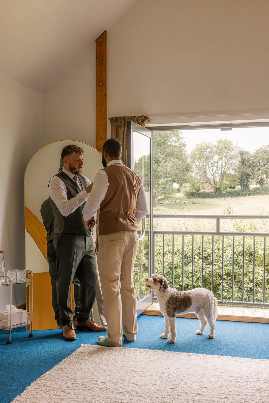 Jack helps Aaron adjust his wedding waistcoat as their dog Milo watches beside the open balcony doors during preparations at Rock Village Hall in Worcestershire, photographed by Just James Weddings.