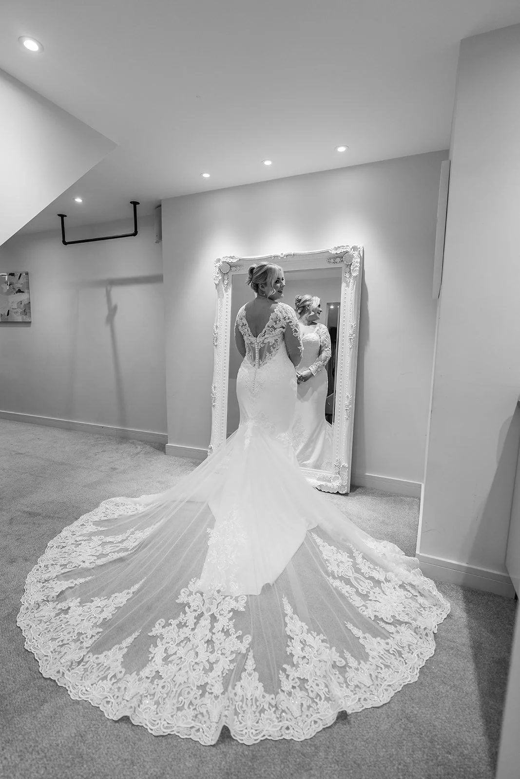 Bride Daisy admiring her lace mermaid wedding dress in a full-length mirror at Andrew & Daisy’s Sandy Cove Hotel wedding in Devon, showcasing the detailed lace train and elegant silhouette during relaxed bridal preparations at this coastal venue.