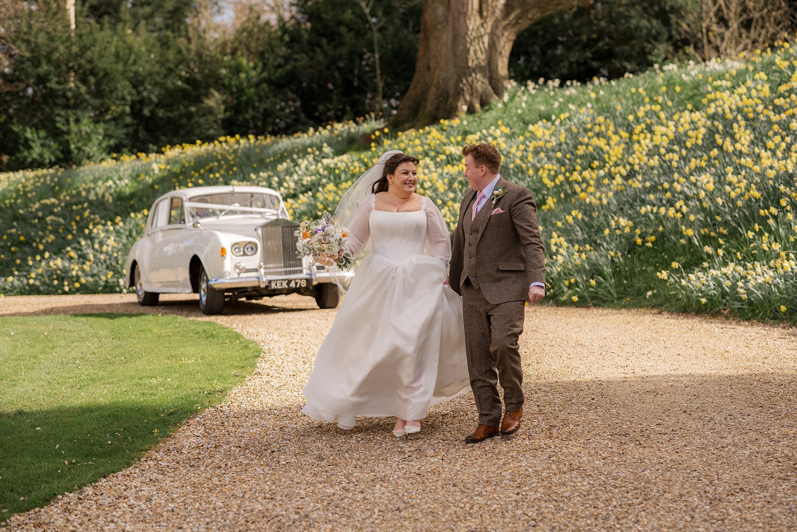 Emily and Josh walk hand in hand through the grounds of Coombe Lodge in Somerset, UK, with a classic wedding car and a hillside of daffodils behind them.
