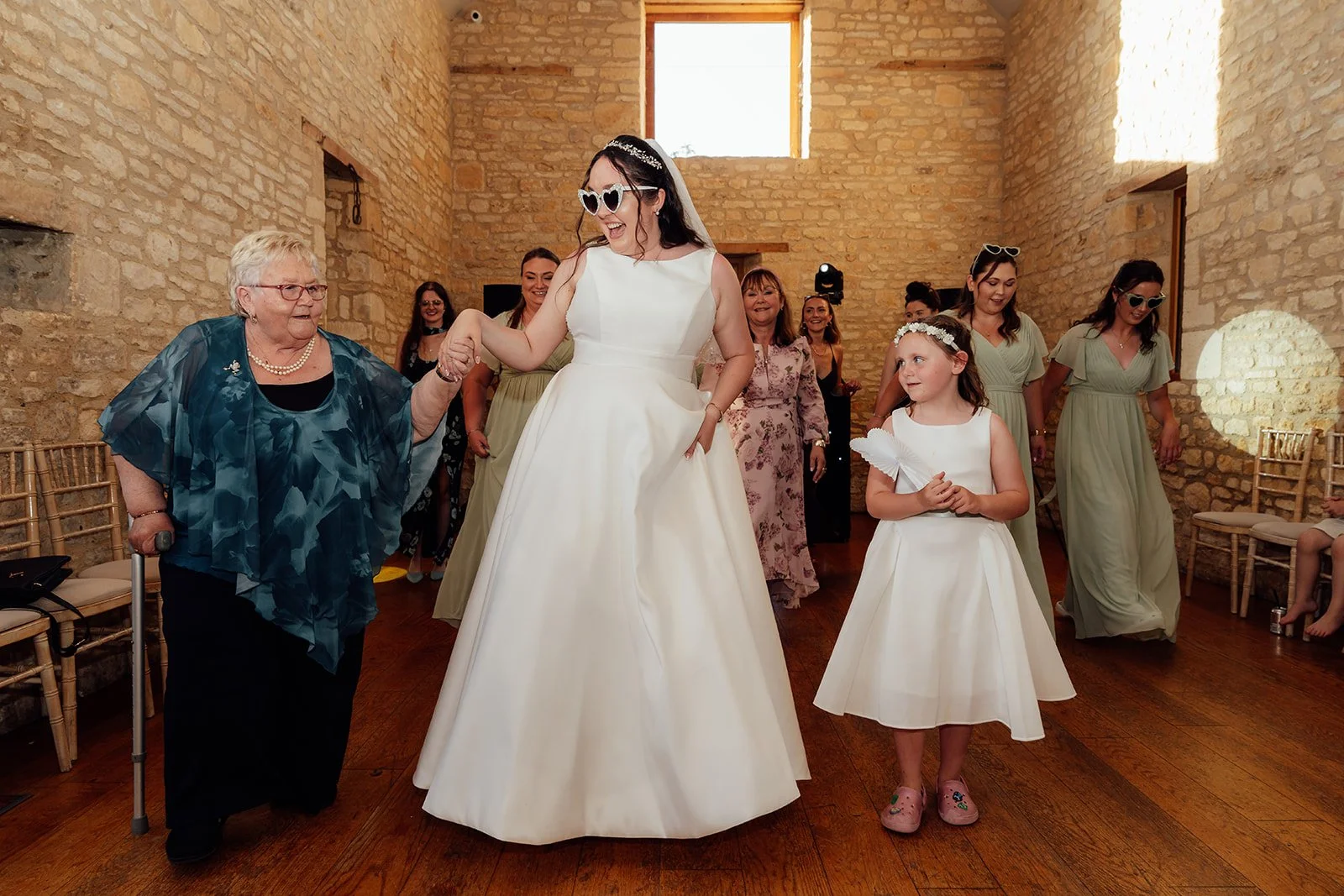 Chloe dances with an older wedding guest and a flower girl during the evening celebration with Armani at The Barn at Upcote in Gloucestershire, photographed by Just James Weddings.