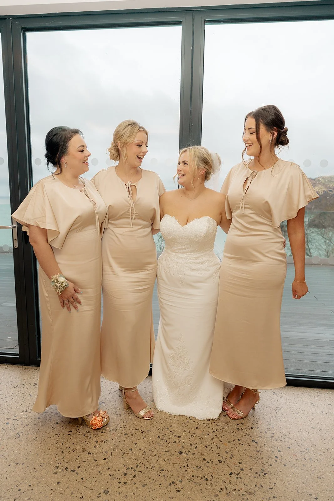 Daisy smiling with her bridesmaids in champagne dresses at Sandy Cove Hotel in Devon, standing before floor-to-ceiling windows with sea views, capturing joyful bridal party moments at this modern coastal wedding venue.