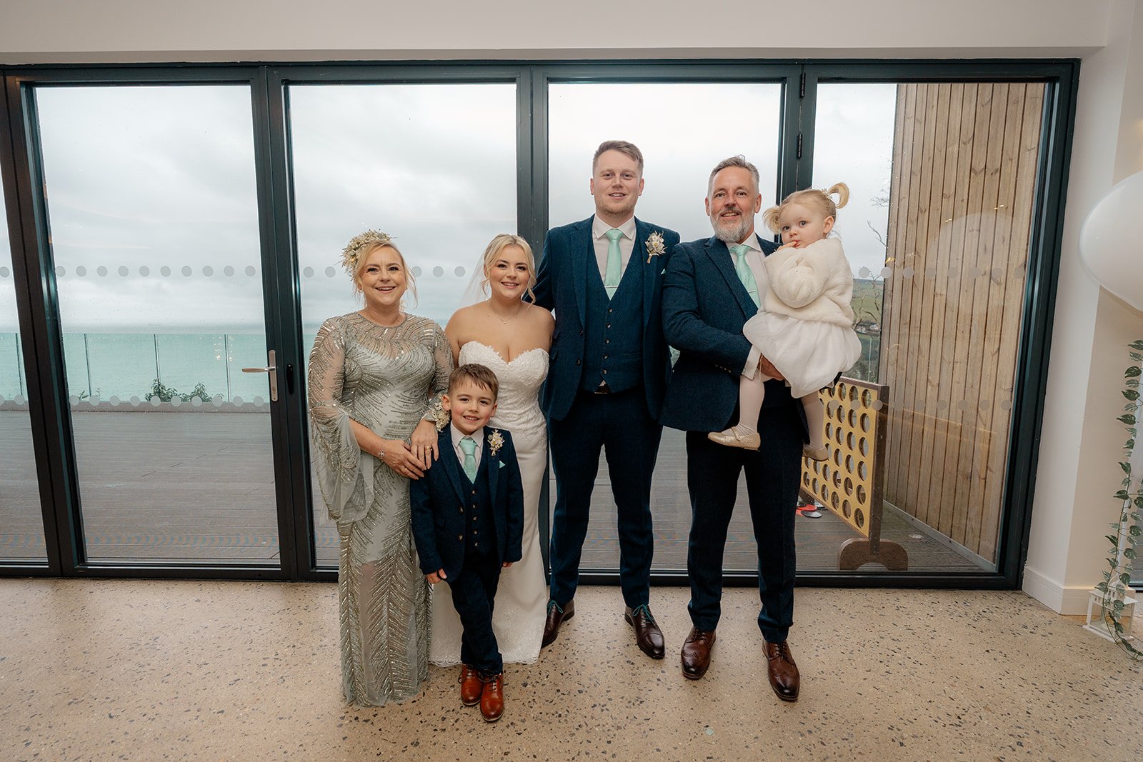Andrew & Daisy posing with close family at Sandy Cove Hotel in Devon, standing before floor-to-ceiling windows with panoramic sea views, capturing a relaxed wedding family portrait with children and coastal backdrop at this Devon seaside venue.
