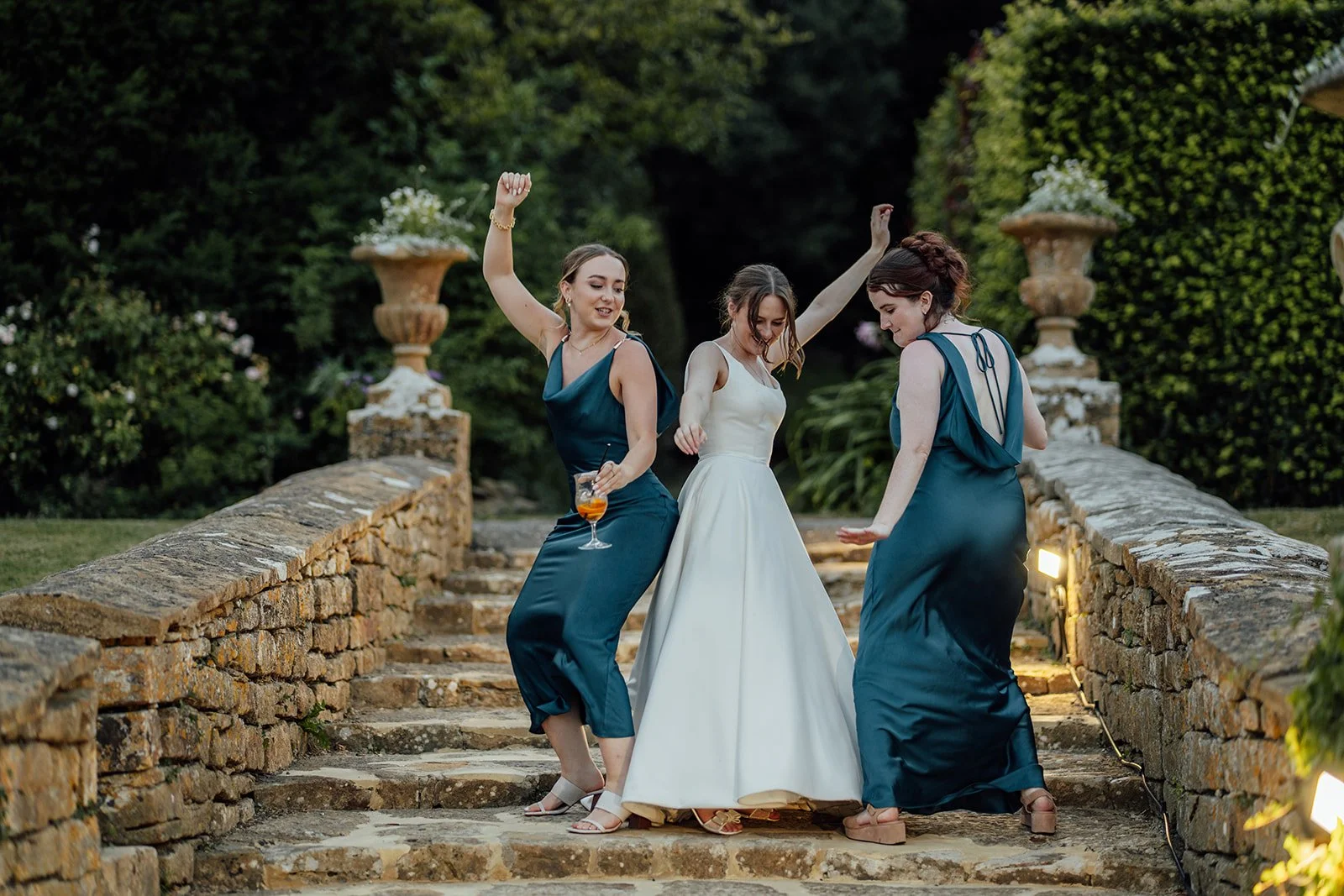 Rachel dances outdoors with two bridesmaids in teal dresses on the stone steps at Brympton House in Somerset, photographed by Just James Weddings.