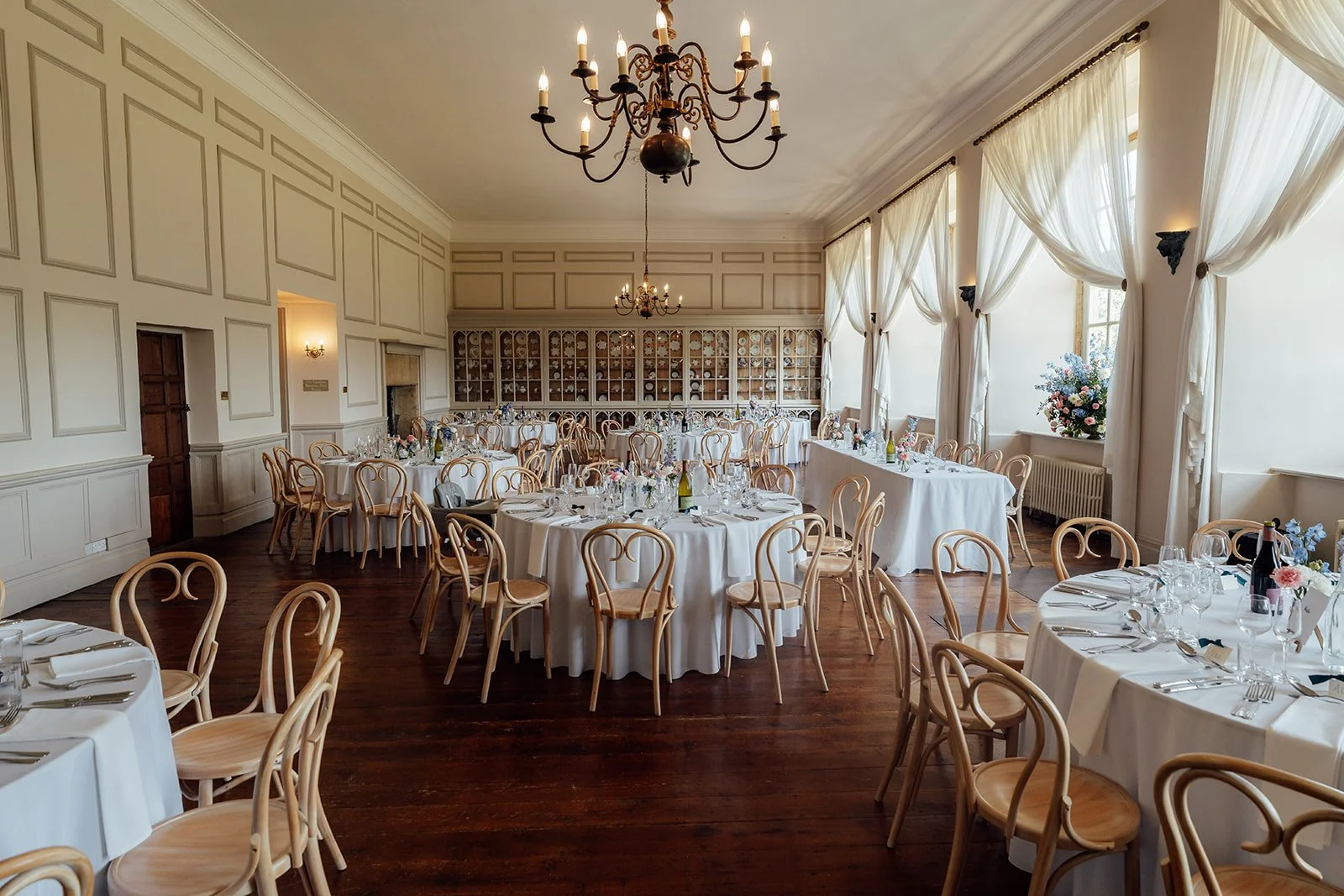 The Ballroom at Brympton House in Somerset, set for a wedding breakfast with round tables, bentwood chairs, chandeliers and tall windows, photographed by Just James Weddings